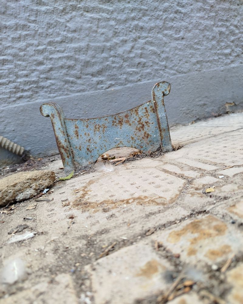 A rusty, pale-blue iron boot scraper — a curved blade between two curled posts — fixed into the pavement at the foot of an old wall in Athens.