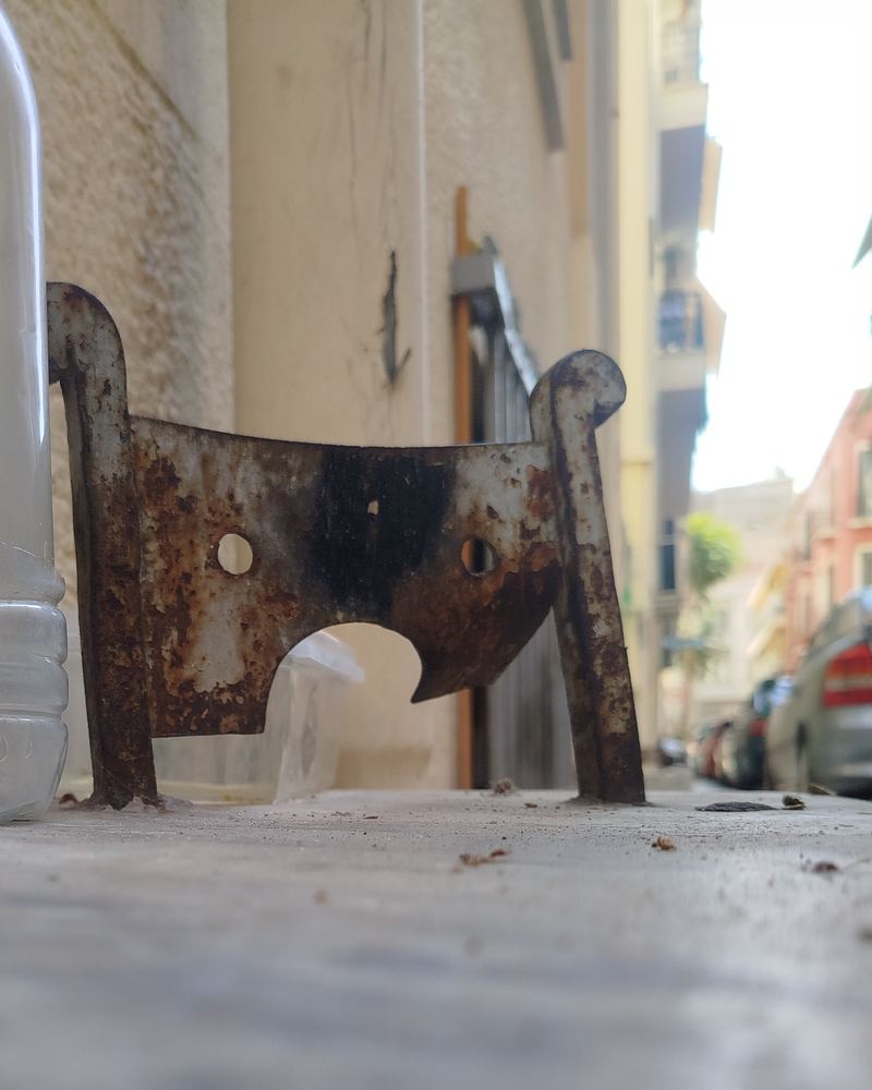 Ground-level view of a rusted iron boot scraper beside a doorway on a narrow Athens street, with parked cars and apartment balconies blurred behind it.